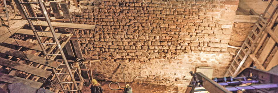 Blick nach Osten auf die Westwand des mehrphasigen Turmes im Ausgrabungsareal im Jahr 1990 unter dem Westsaal. Die Verputzreste im unteren Abschnitt kennzeichnen einen Teil des ersten, lang rechteckigen Turmes (11. Jahrhundert). Vorne im Bild rechts und links zwei sog. Dachdeckeraufzüge zum Materialtransport. – Foto: S. Wielonek, WiBA e.V.