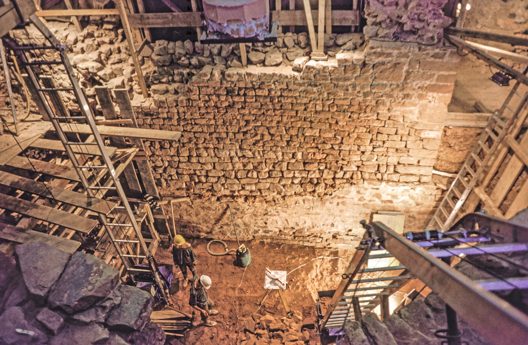 Blick nach Osten auf die Westwand des mehrphasigen Turmes im Ausgrabungsareal im Jahr 1990 unter dem Westsaal. Die Verputzreste im unteren Abschnitt kennzeichnen einen Teil des ersten, lang rechteckigen Turmes (11. Jahrhundert). Vorne im Bild rechts und links zwei sog. Dachdeckeraufzüge zum Materialtransport. – Foto: S. Wielonek, WiBA e.V.